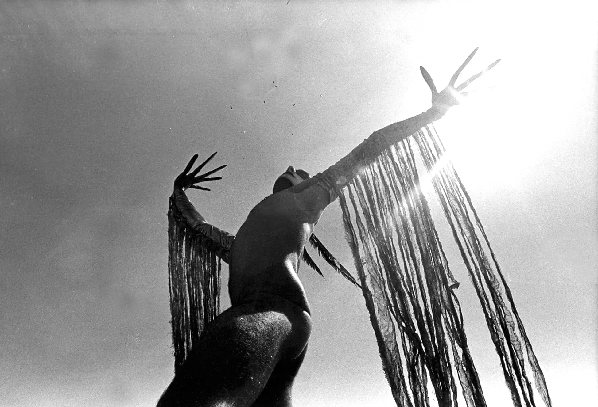 Obra de Laercio Redondo: fotografia em preto e branco de uma figura de braços abertos contra o céu, com longas franjas presas aos braços